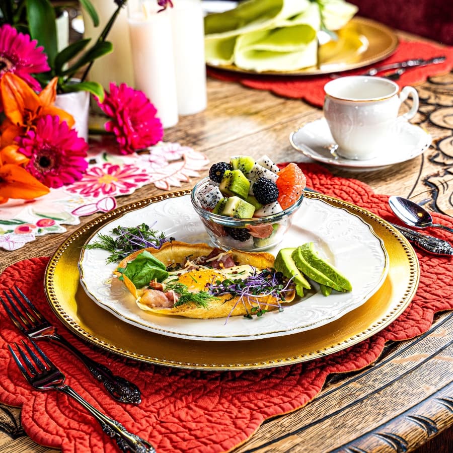 A beautifully arranged breakfast plate featuring an omelet with greens, avocado, and a fruit salad in a bowl, set on a vibrant table.