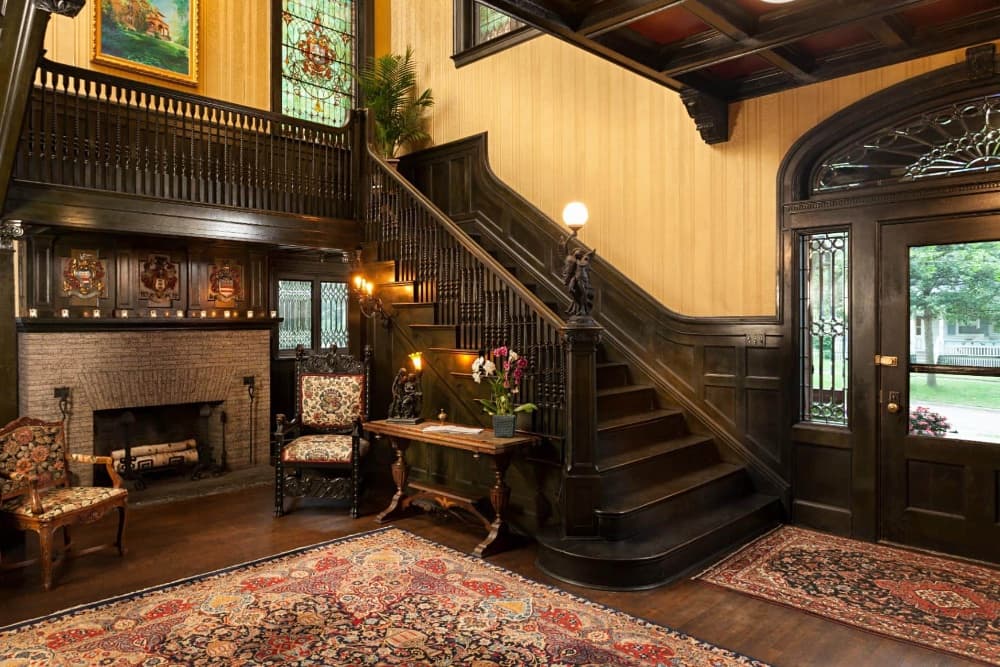 A richly decorated foyer featuring a grand staircase, fireplace, and vintage furnishings.