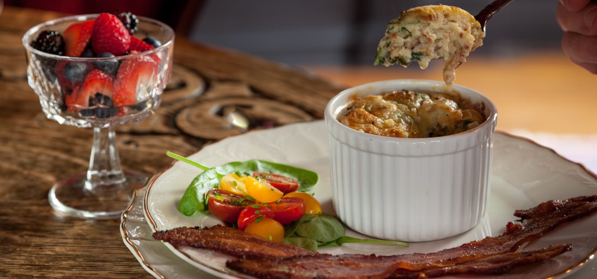 A plated breakfast featuring a ramekin of baked dish, crispy bacon, colorful cherry tomatoes, spinach, and a glass of mixed berries.