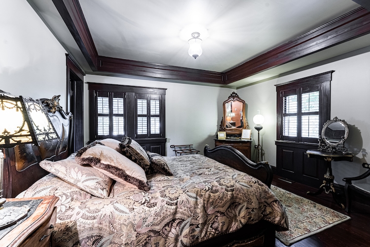 Luxurious vintage-style bedroom with dark wood trim, a carved wooden bed frame, and patterned bedding, a vanity table with mirror, a writing desk, and an ornate dresser.