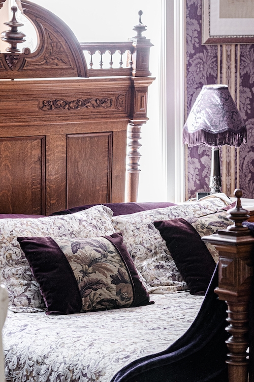 Close up of sumptuous queen size carved oak bed, plump pillows, and nightstand with boudoir lamp
