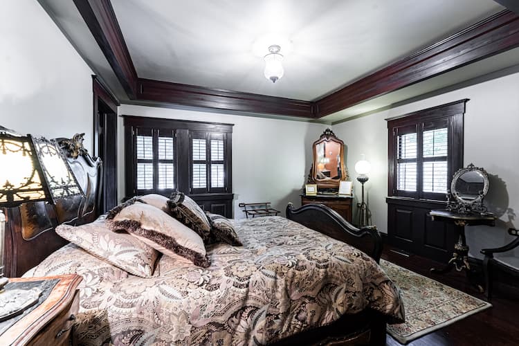 Luxurious vintage-style bedroom with dark wood trim, a carved wooden bed frame, and patterned bedding, a vanity table with mirror, a writing desk, and an ornate dresser.