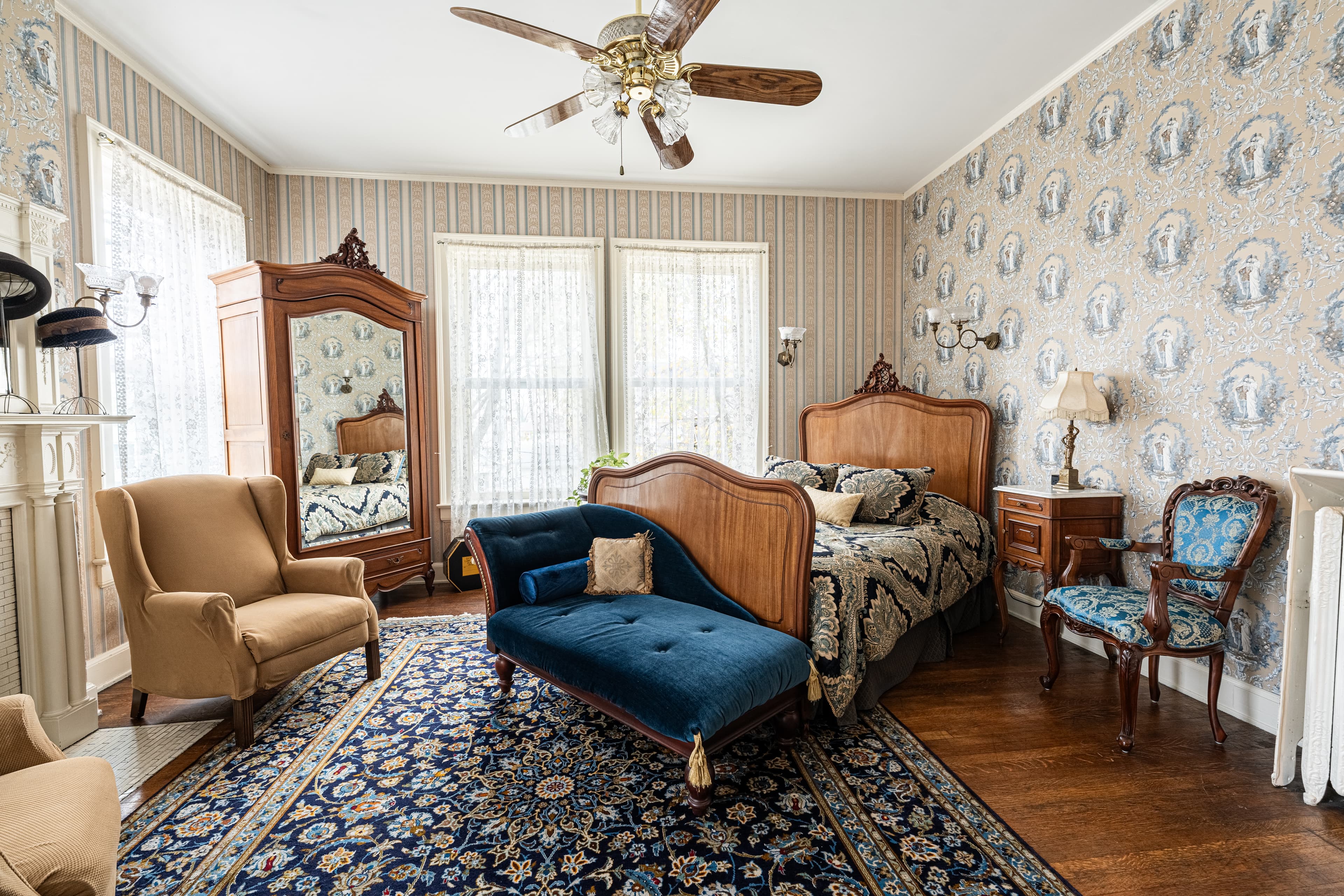 Elegant vintage-style bedroom with a queen bed, ornate wood headboard, blue velvet chaise lounge,and floral wallpaper.