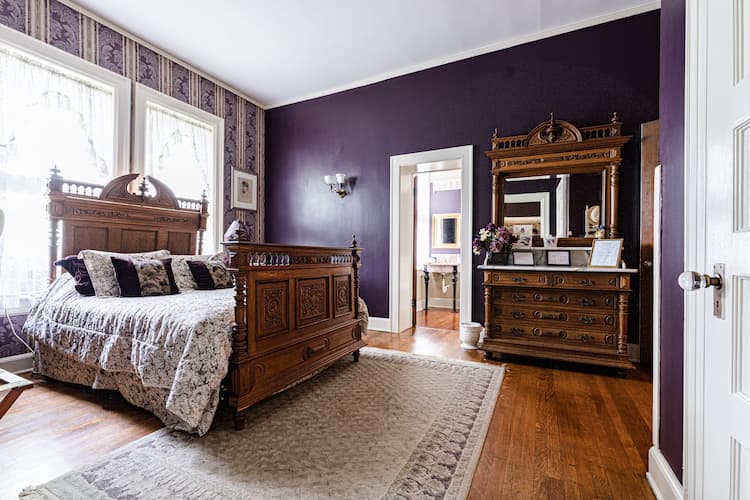 View of Letitia's Bedroom with queen size bed in front of west facing twin windows . Dark eggplant walls with huge tapestry.
