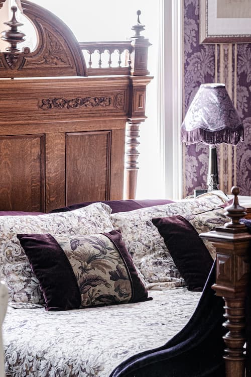 Close up of sumptuous queen size carved oak bed, plump pillows, and nightstand with boudoir lamp