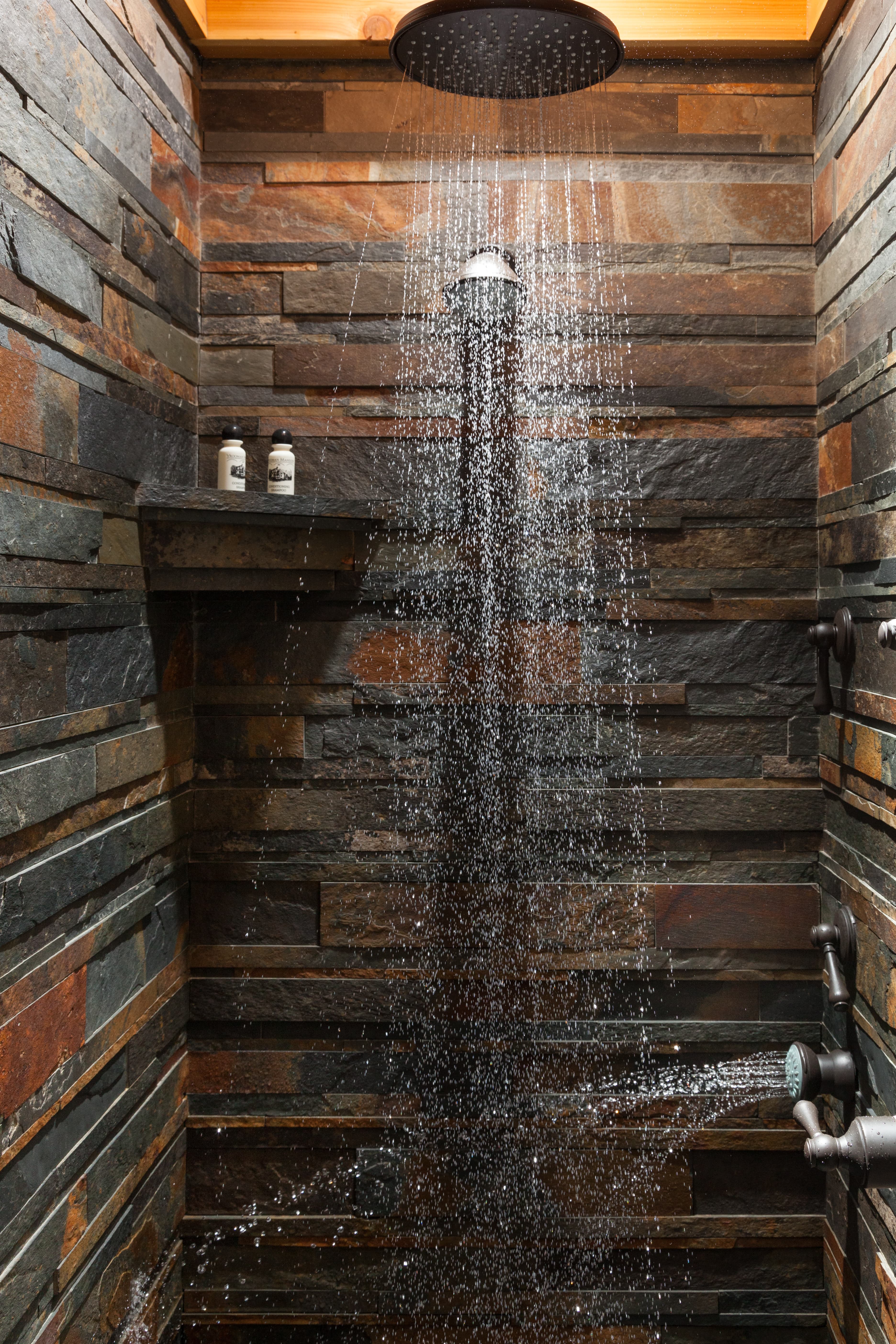 Modern walk-in shower with a ceiling-mounted rainfall showerhead and textured stacked stone walls in earthy tones.