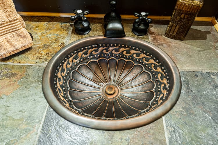An ornate, oval-shaped copper sink with intricately patterned basin installed in a countertop made of gray and brown stone tiles.