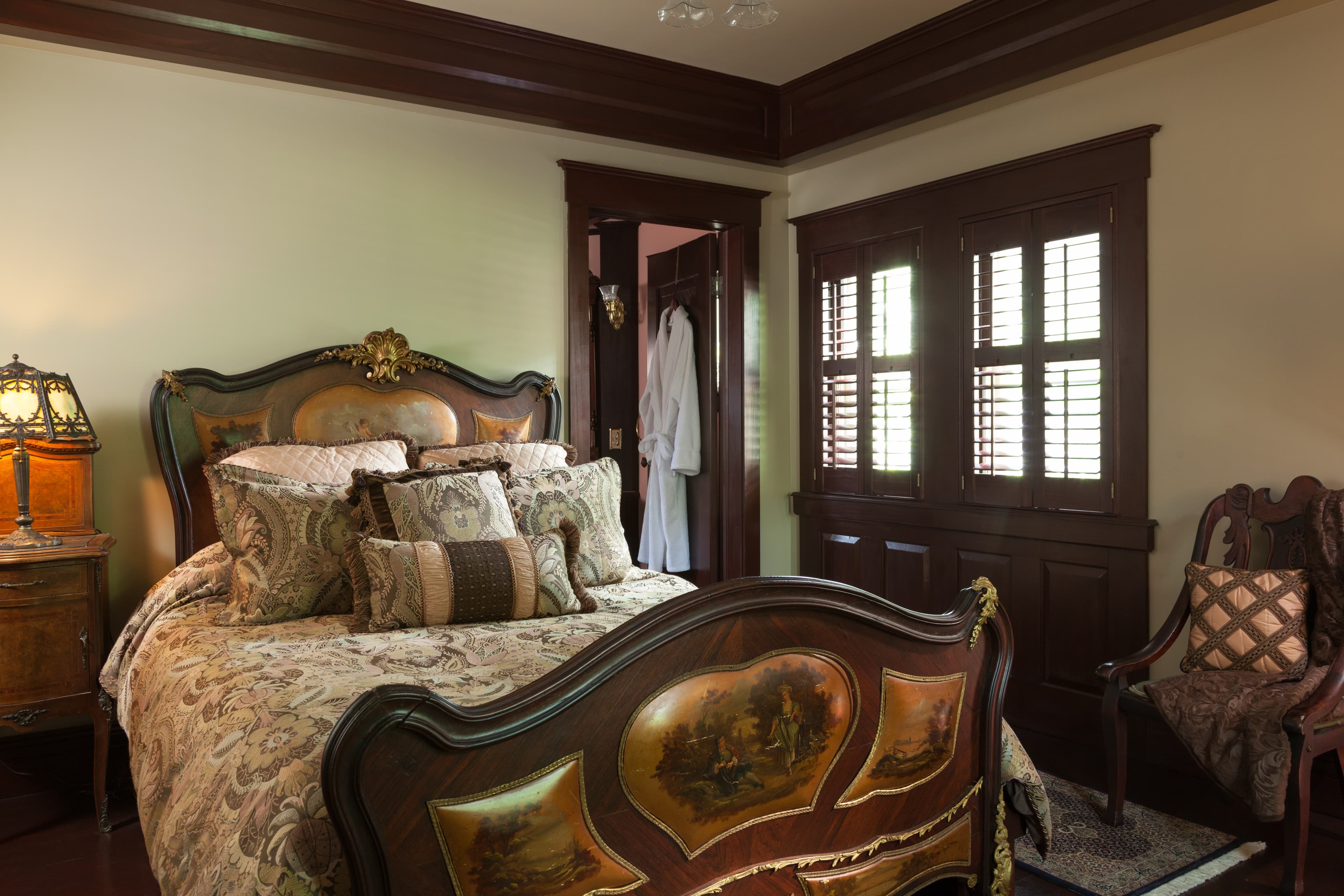A warmly lit bedroom featuring a large, ornate wooden bed with a curved headboard and footboard and a wooden nightstand with a stained-glass lamp beside the bed.