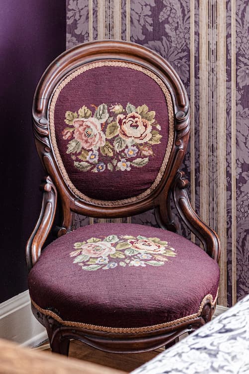 Purple antique chair with needlepointed floral seat and back in Letitia's room next to the bed