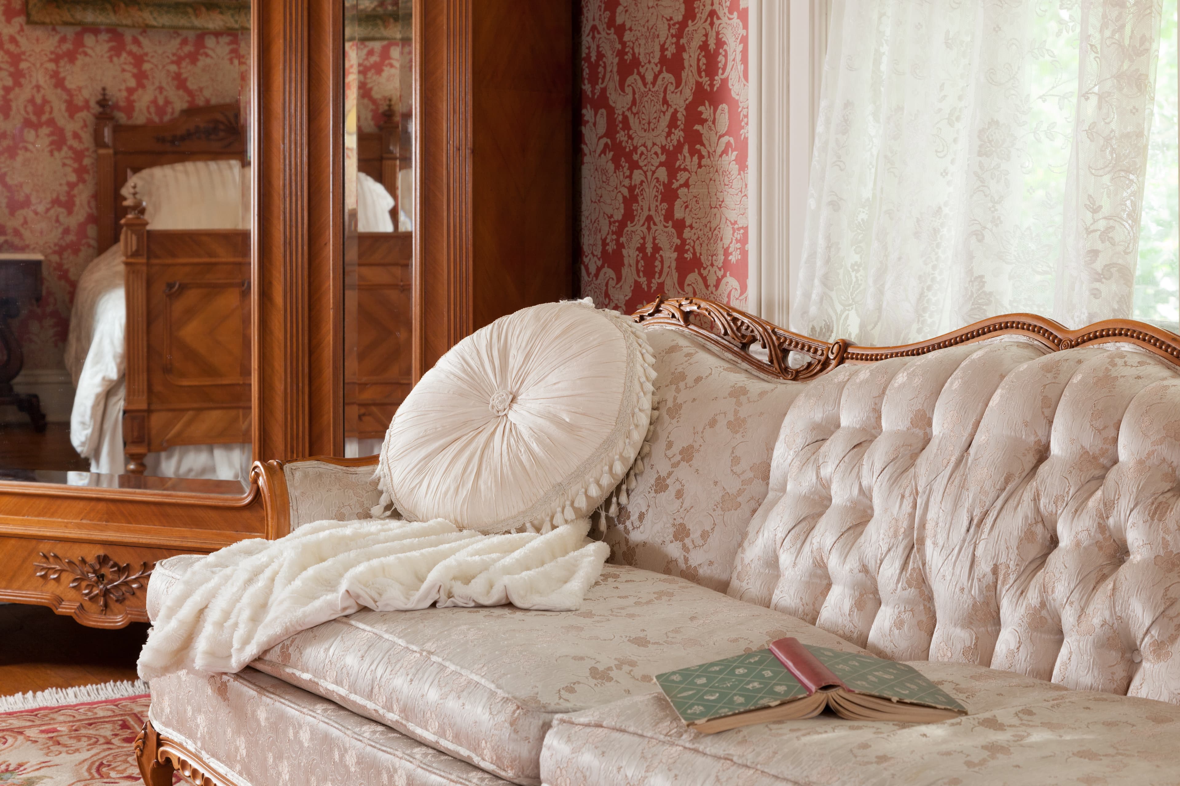 A softly lit interior space features a plush, light-colored chaise lounge and a large mirrored armoire reflecting the antique queen size bed.