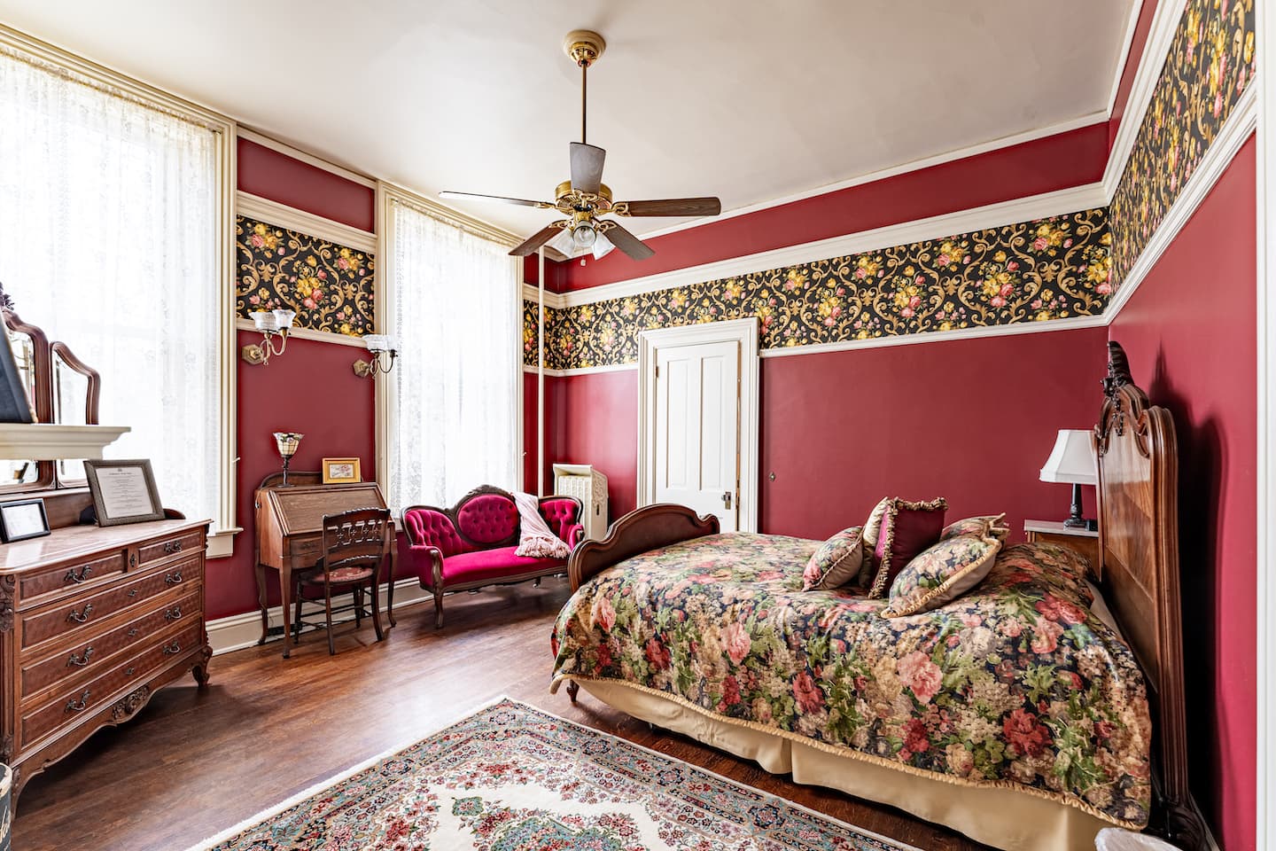 A warmly lit bedroom with red walls, floral wallpaper trim, and antique wooden furniture including a large bed with a floral comforter.