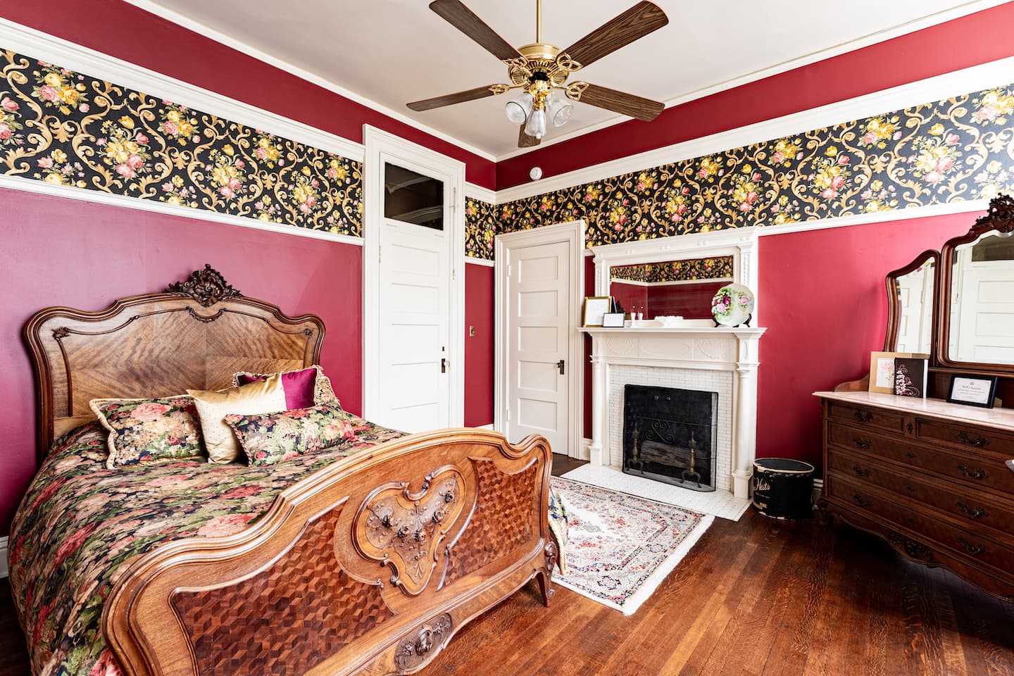 Bedroom with deep red walls accented by a decorative floral wallpaper border near the ceiling, a queen antique wooden bed with an intricately carved headboard and footboard, and a decorative fireplace.