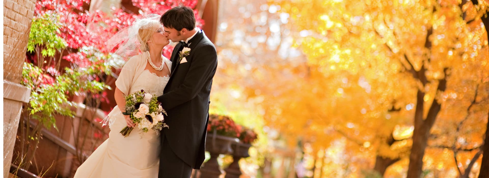 A bride and groom kiss amidst vibrant autumn foliage.