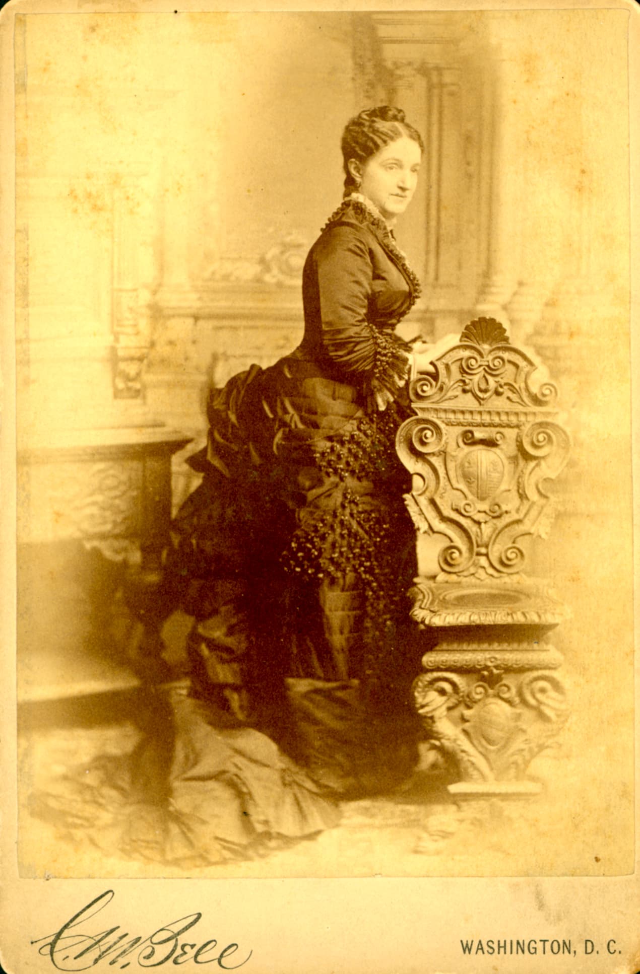 A woman in a formal 19th-century dress poses beside an ornate chair in a vintage photograph.