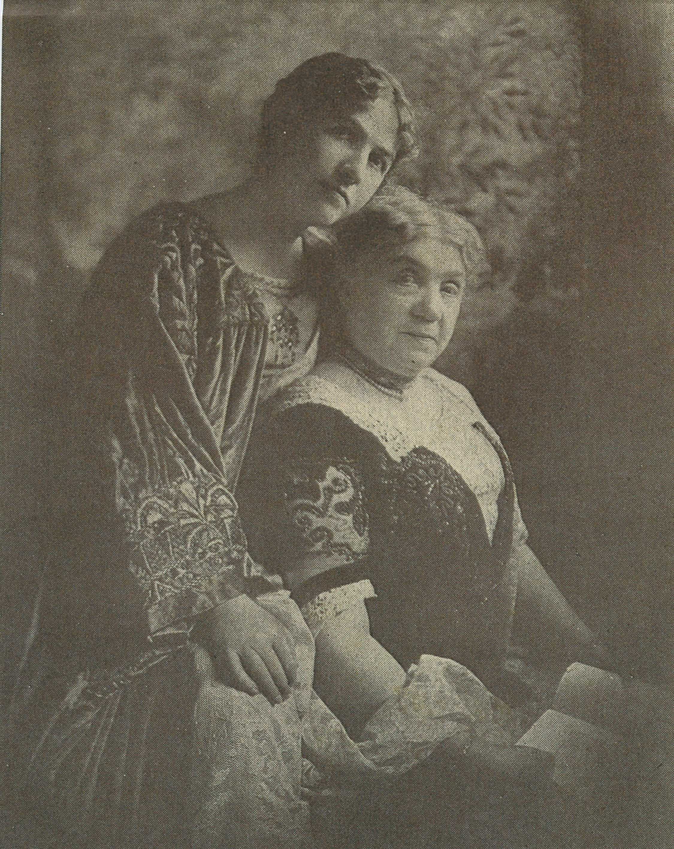 Two women pose closely together, one seated and the other standing behind her, both dressed in vintage clothing.