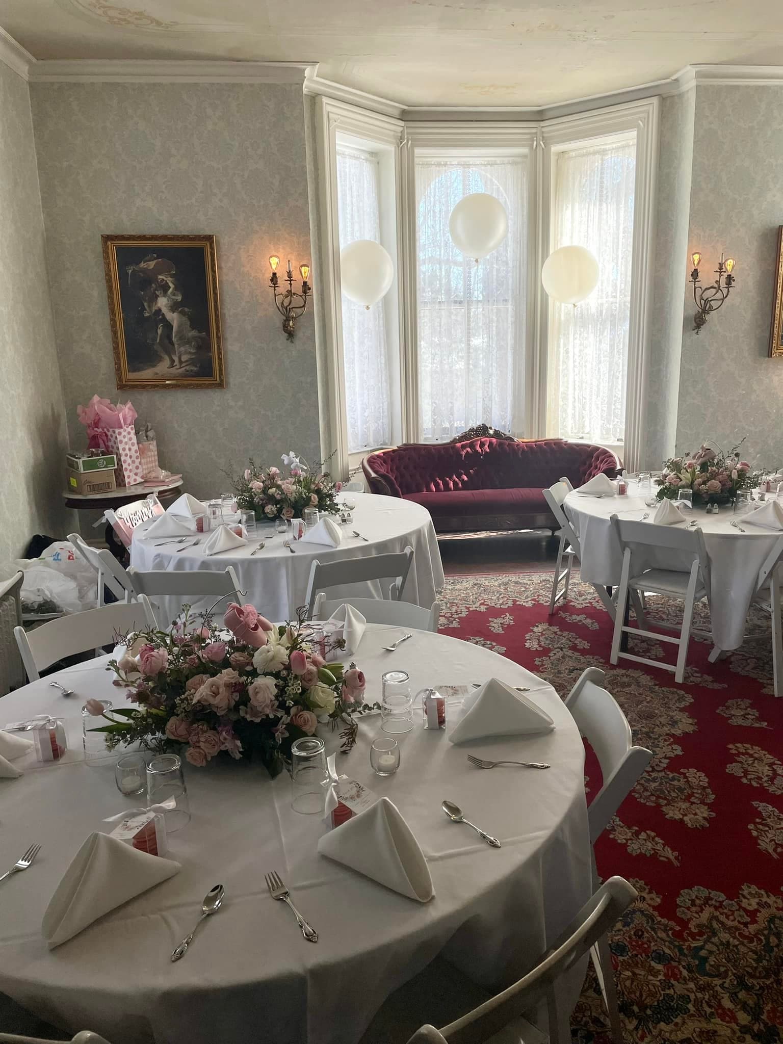 Elegant dining room setup with tables adorned with floral centerpieces and white napkins, featuring a cozy seating area and decorative balloons.