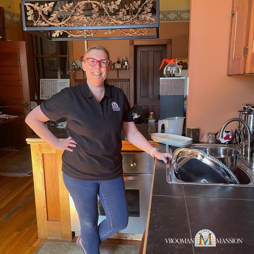A smiling woman in a black polo shirt stands by a kitchen counter with a sink and various kitchen items.