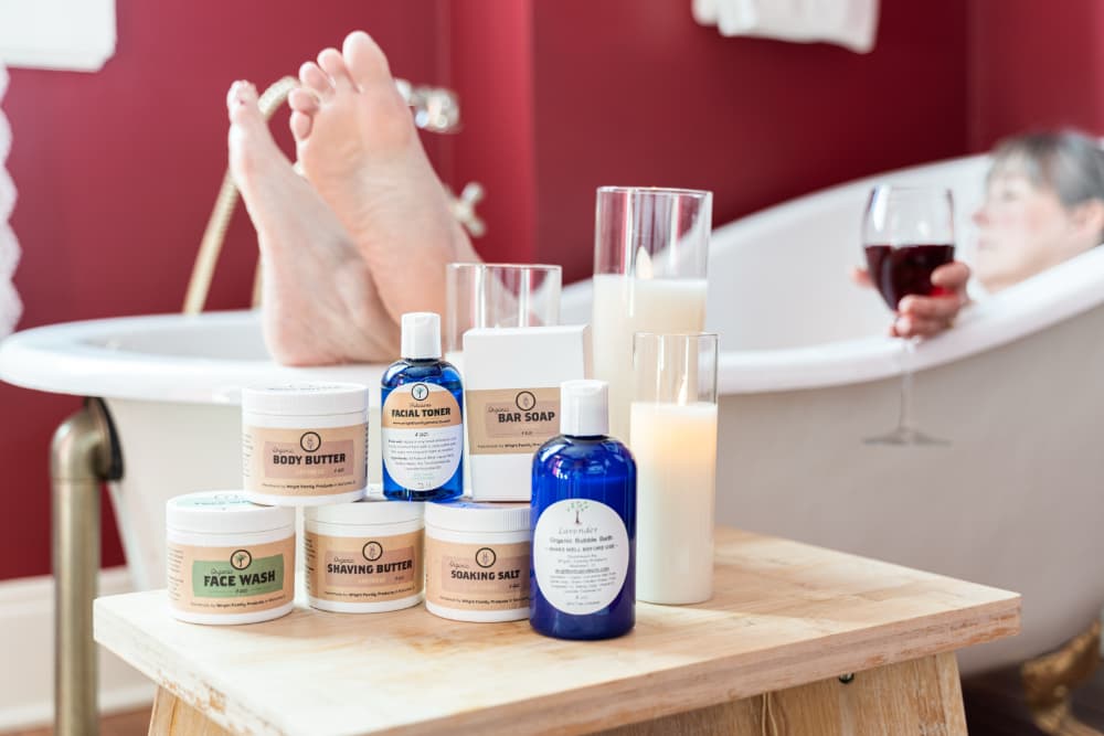 A person relaxes in a bathtub while surrounded by various skincare products and candles.