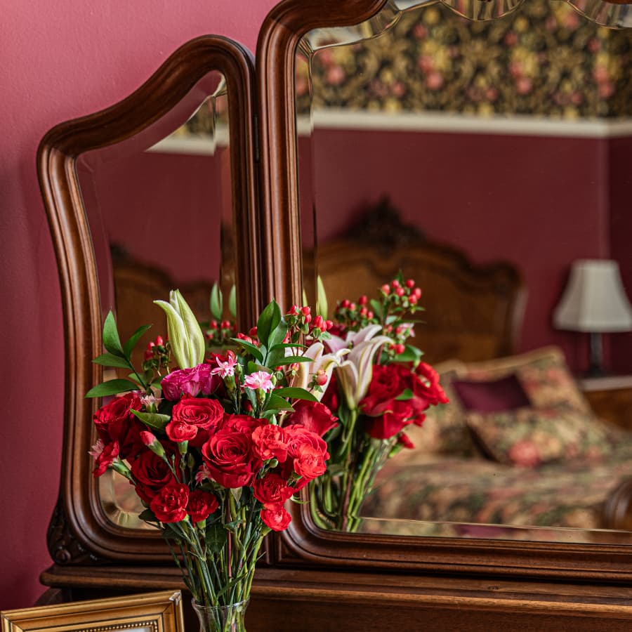 A vibrant bouquet of red and pink flowers reflects in a wooden mirror against a pink wall.