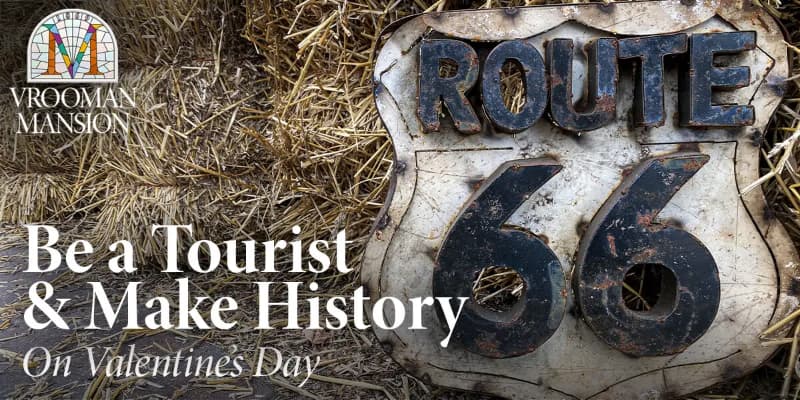 A weathered Route 66 sign with the message "Be a Tourist & Make History On Valentine's Day" beside hay.
