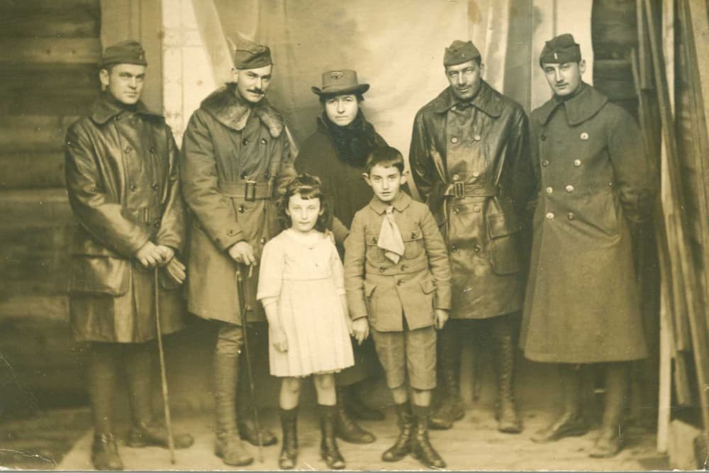 A group of six adults and two children poses together in vintage clothing, likely from the early 20th century.