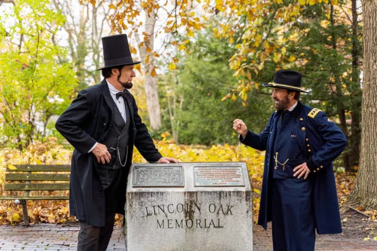Two men in period costumes stand by the Lincoln Oak Memorial, surrounded by autumn foliage.