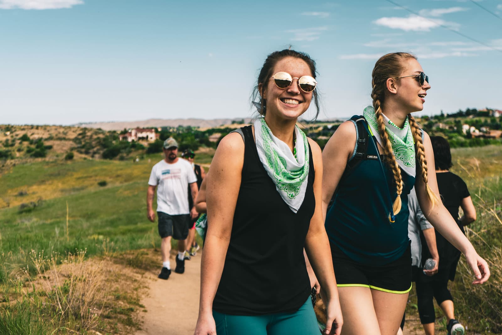 Two women in athletic gear and sunglasses smile while hiking on a scenic trail, with others in the background. Two women in athletic gear and sunglasses smile while hiking on a scenic trail, with others in the background.