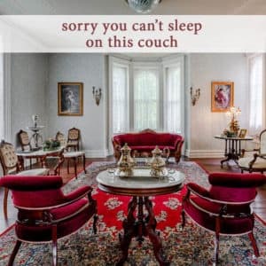 A stylish parlor with red upholstered furniture and elegant décor, featuring a sign that reads "sorry you can't sleep on this couch."