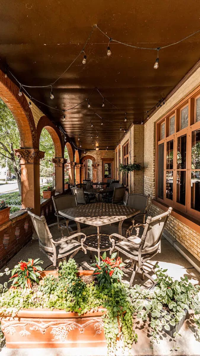 A cozy outdoor dining area with metal chairs, a round table, and potted plants under a covered patio.