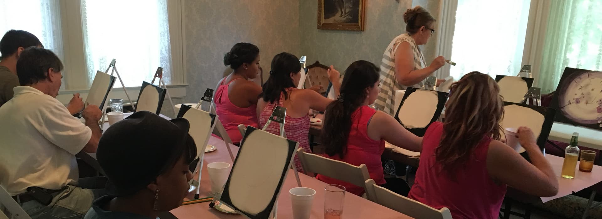 People participating in a painting class, seated at tables with easels and supplies.