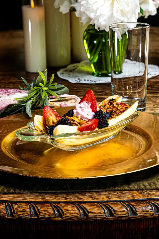 A glass dish of banana split topped with strawberries, blackberries, and whipped cream on a golden platter.