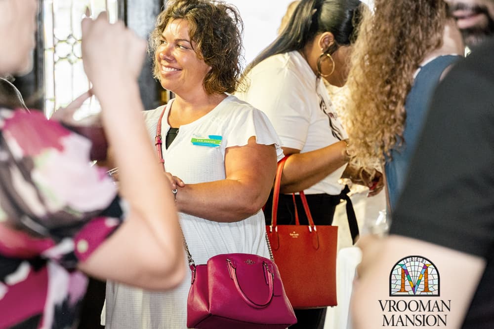 A smiling woman in a white dress with a pink handbag engages with others at an event.