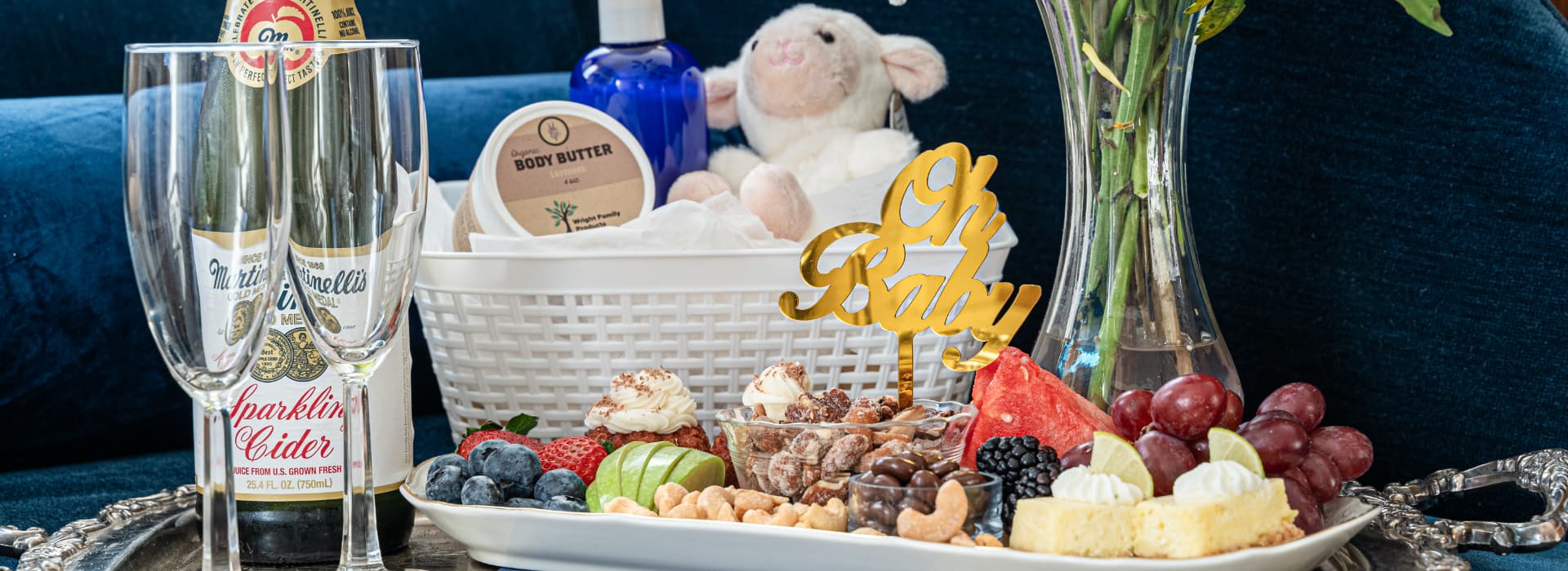 A festive arrangement featuring sparkling cider, a variety of fruits, desserts, and a "Oh Baby" cake topper, set on a decorative tray.