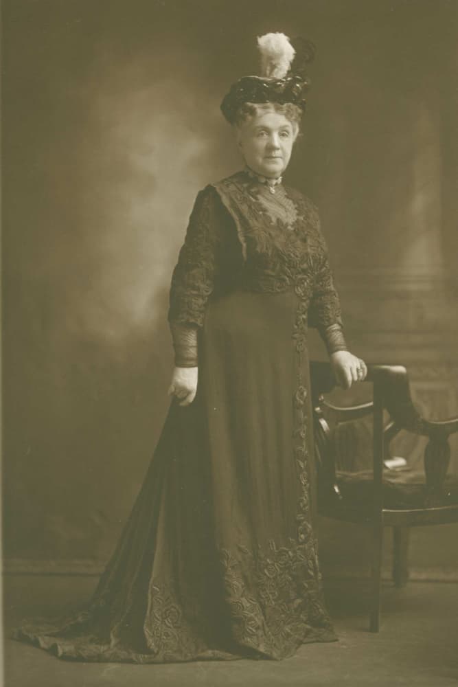 A woman in a formal, intricately embroidered dark dress with a feathered hat poses beside a chair in a vintage photograph.