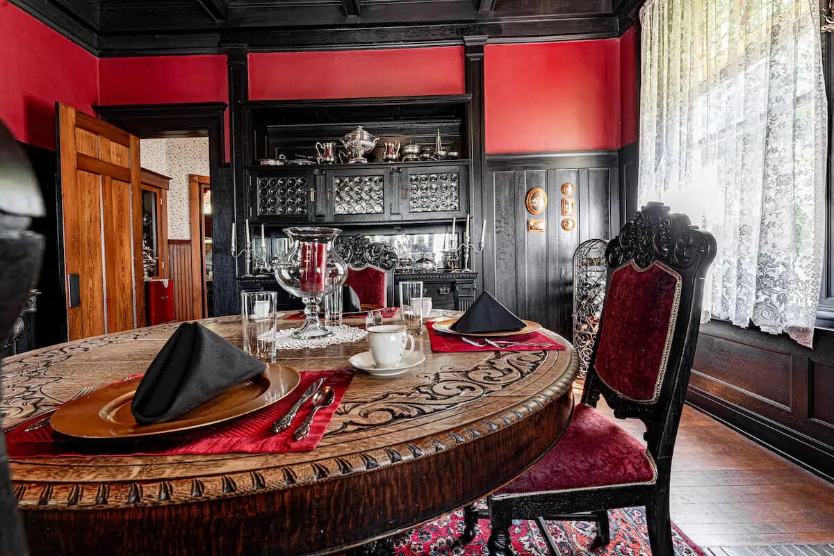 A richly decorated dining room featuring a round wooden table set with elegant tableware against a backdrop of dark wood and red walls.