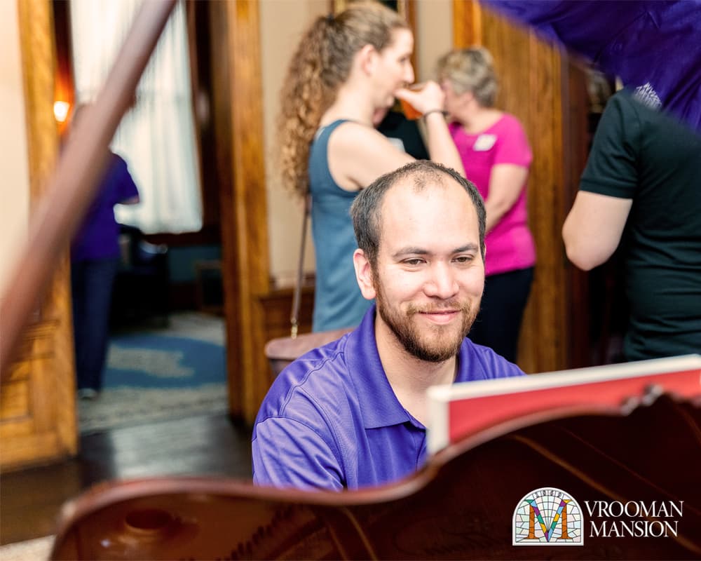 A man in a purple shirt smiles while playing the piano, surrounded by people in a warmly lit room.