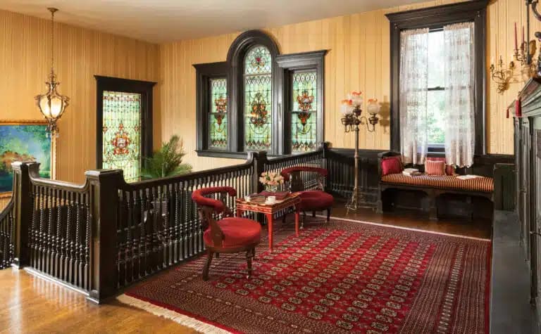 A cozy room featuring dark wood accents, stained glass windows, and red upholstered chairs on a decorative rug.