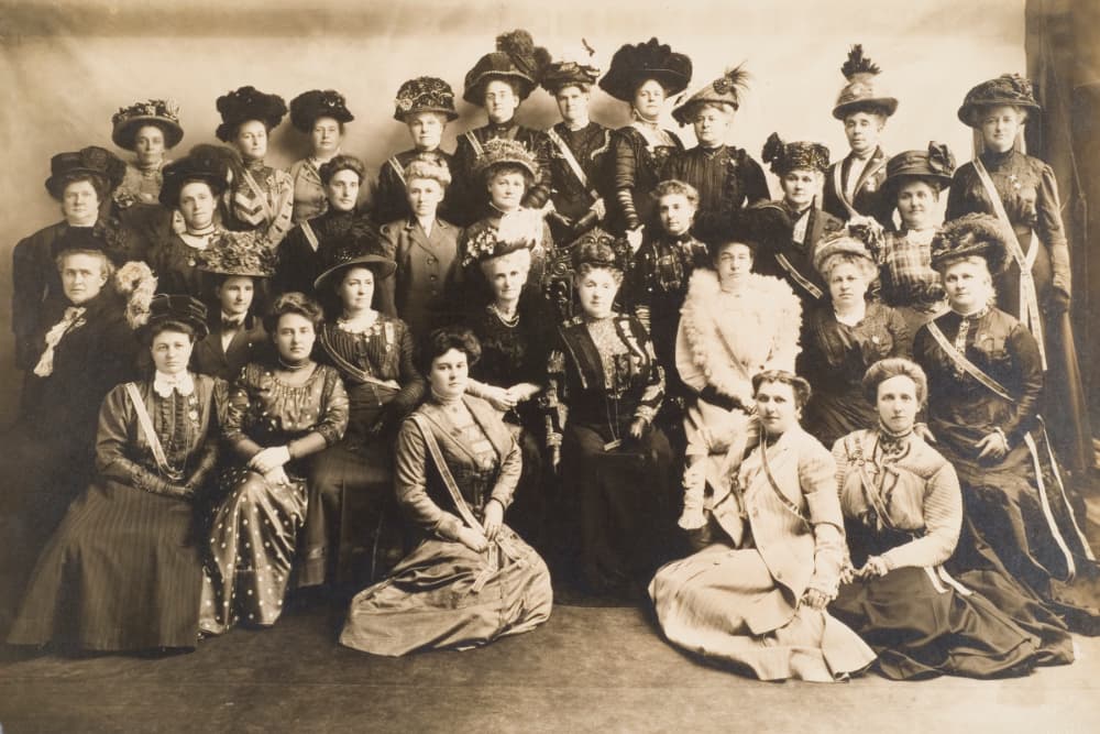 A historic black-and-white photograph featuring a large group of women in elaborate dresses and hats, posing together.