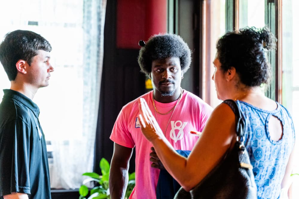 Three people engage in conversation indoors, with one man in a pink shirt attentively listening.