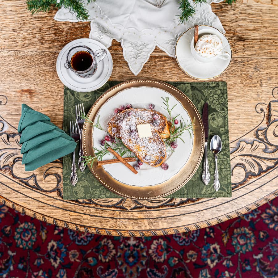 A beautifully arranged breakfast plate with French toast, garnished with berries and herbs, beside a cup of coffee and a festive napkin on a wooden table.