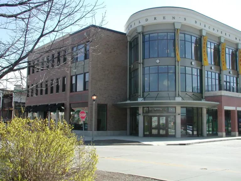 A modern building with large windows labeled "Children's Discovery Museum" next to a small brick structure. A modern building with large windows labeled "Children's Discovery Museum" next to a small brick structure.