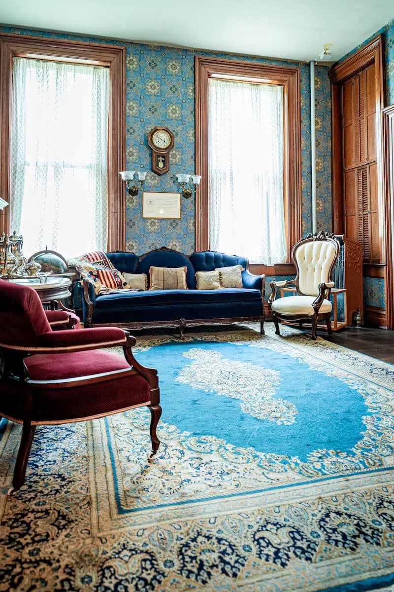 A vintage living room featuring a blue sofa, a white armchair, and decorative wallpaper.