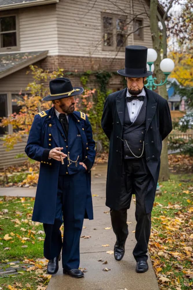 Two men in 19th-century attire walk together on a sidewalk surrounded by autumn leaves.
