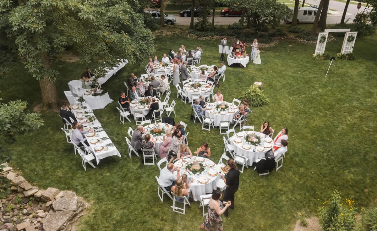 Aerial view of a large outdoor gathering with guests seated at round tables, enjoying a meal in a grassy area.