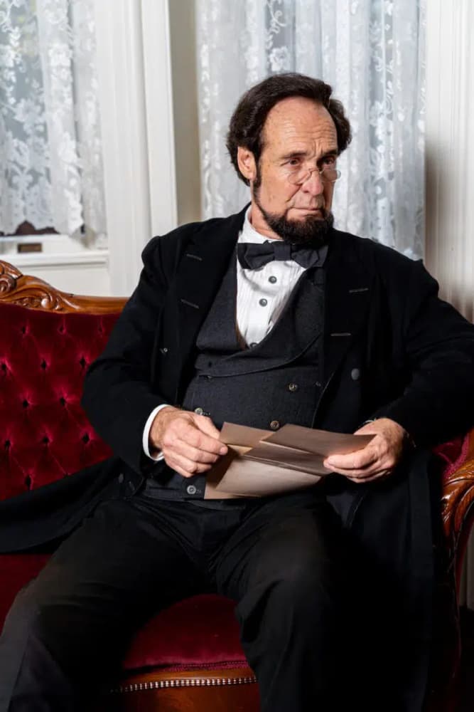 A man dressed in 19th-century clothing sits on a plush chair, holding a letter.