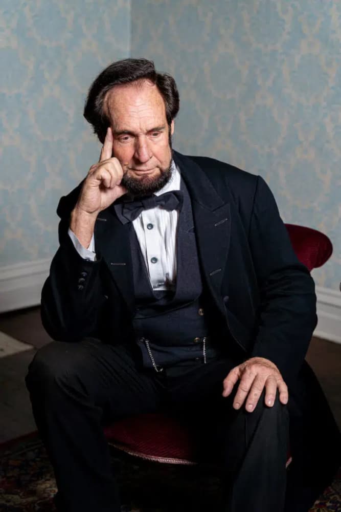 A man in a formal black suit sits pensively in a chair against a patterned wall.