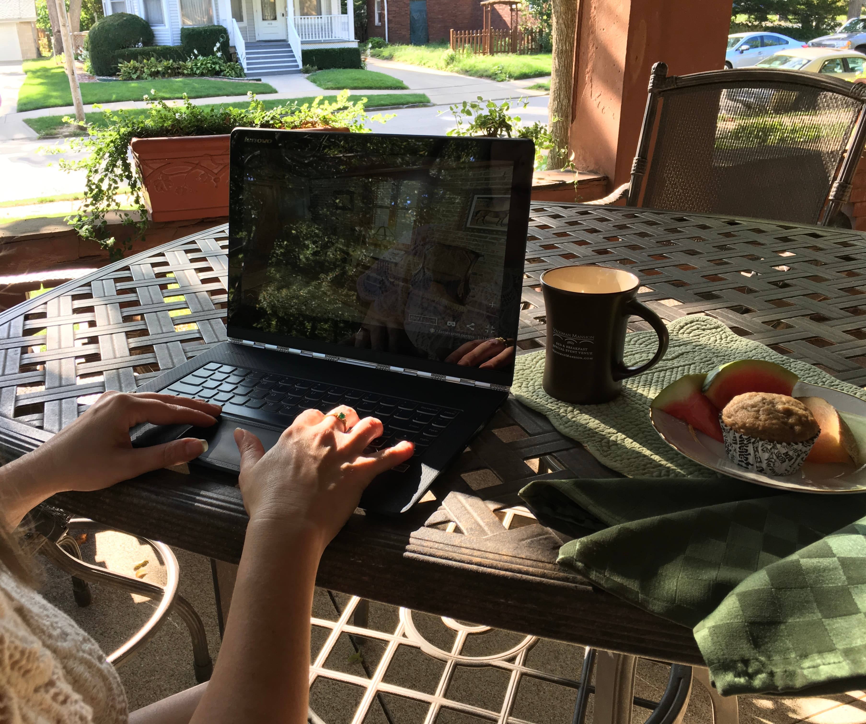 A person is working on a laptop at a table with a cup of coffee, fruit, and a muffin in a sunlit outdoor setting.