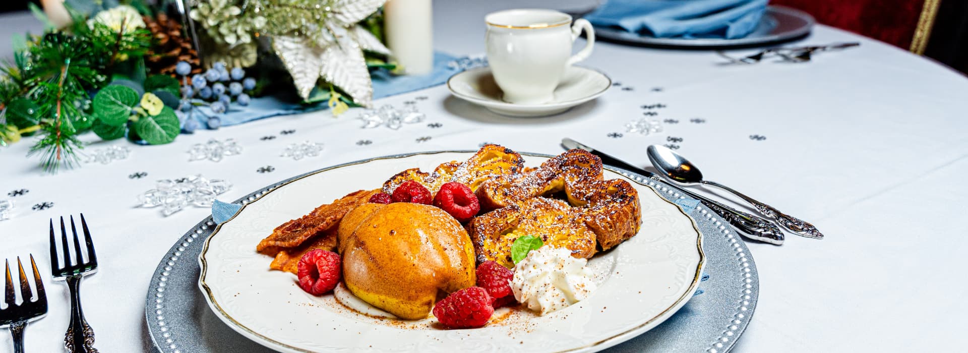 A beautifully arranged breakfast plate featuring French toast, fresh raspberries, and a pear slice, set on an elegant table with decorative greenery.