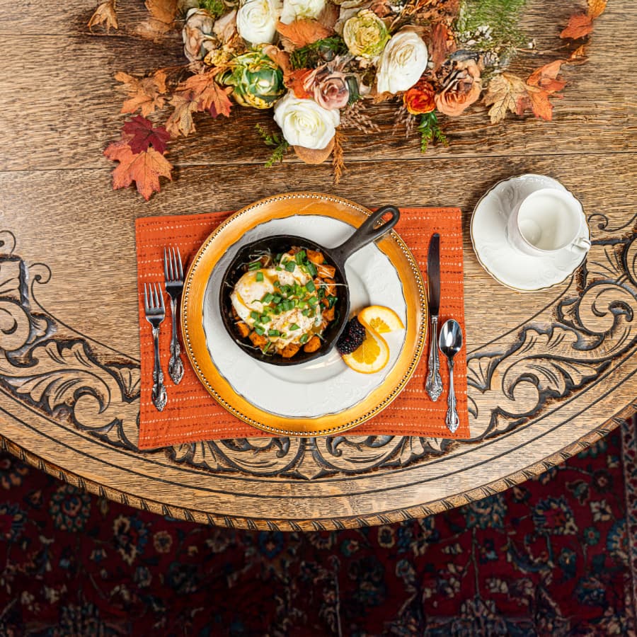 A beautifully arranged table setting featuring a skillet dish garnished with green onions, an orange slice, and a side of blackberries, alongside a cup and saucer.