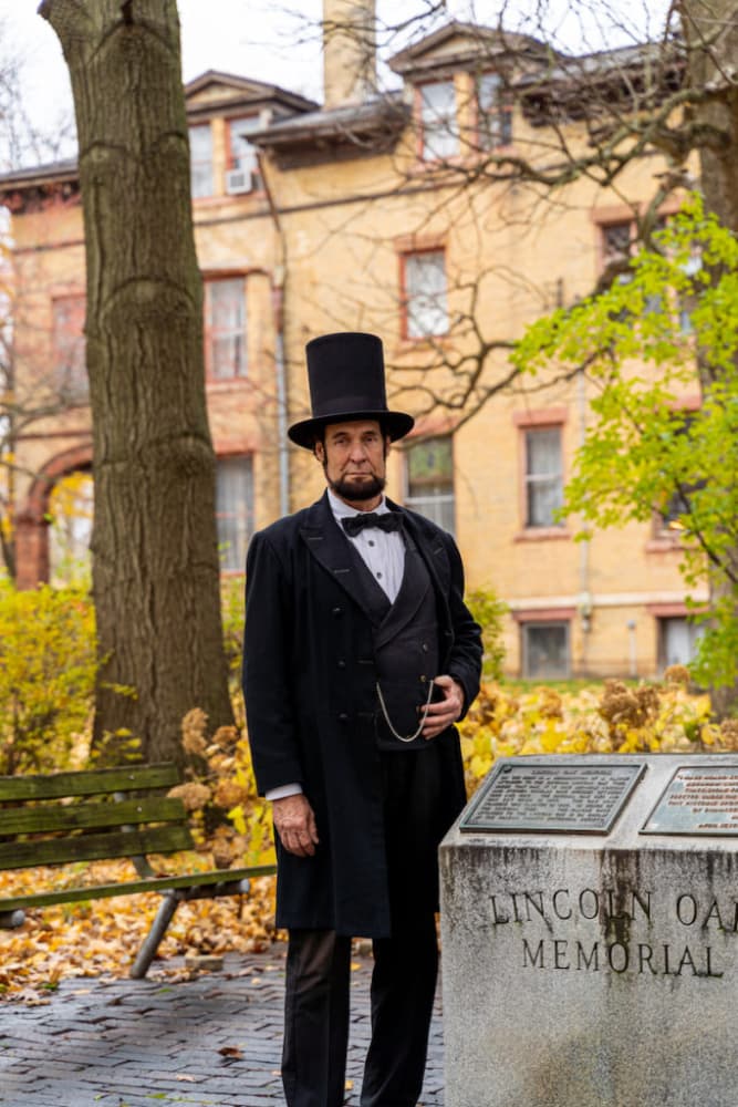 A man dressed as Abraham Lincoln stands beside the Lincoln Oak Memorial in a park during autumn.